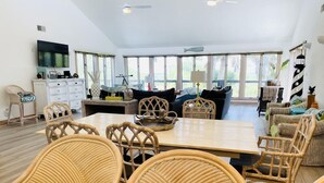 A spacious living room with a large dining table, wicker chairs, a sofa, and a TV mounted on the wall.