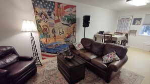 Ein Wohnzimmer mit brauner Ledersofa, einem Couchtisch, einer Lampe, einem Wandbild von Route 66 und einem Essbereich im Hintergrund.