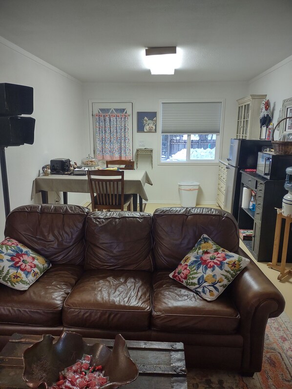 A living room with a brown leather sofa, a dining table with chairs, a kitchen area with appliances, and a window with a view of the outside. - Private Cottage to nurture the soul (MIRAMONTE)