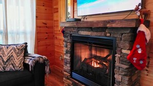 L’intérieur chaleureux d’un chalet avec une cheminée en pierre, un téléviseur fixé au-dessus et une chaussette de Noël accrochée à la cheminée.