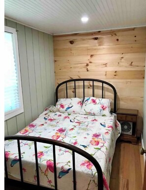 Un lit simple avec une couvre-lit à motifs floraux, dans une pièce aux murs en bois et dotée d’une fenêtre.