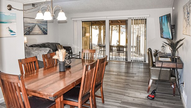 A dining area with a wooden table and chairs, a chandelier, and a view through a glass door to an outdoor patio.