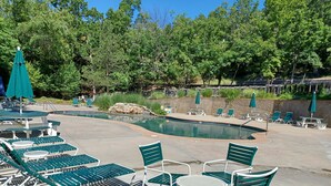 A pool area with green lounge chairs and tables, surrounded by trees.