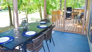 An outdoor dining area with a covered patio, a table set for two with blue plates and cutlery, wicker chairs, and a grill in the background.