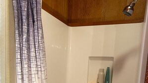Área de chuveiro com parede revestida de azulejos brancos, chuveiro e prateleira com produtos de higiene pessoal.