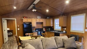 Interior acolhedor de uma cabana com teto de madeira, cozinha compacta, área de refeições com sofá e janela com persianas.