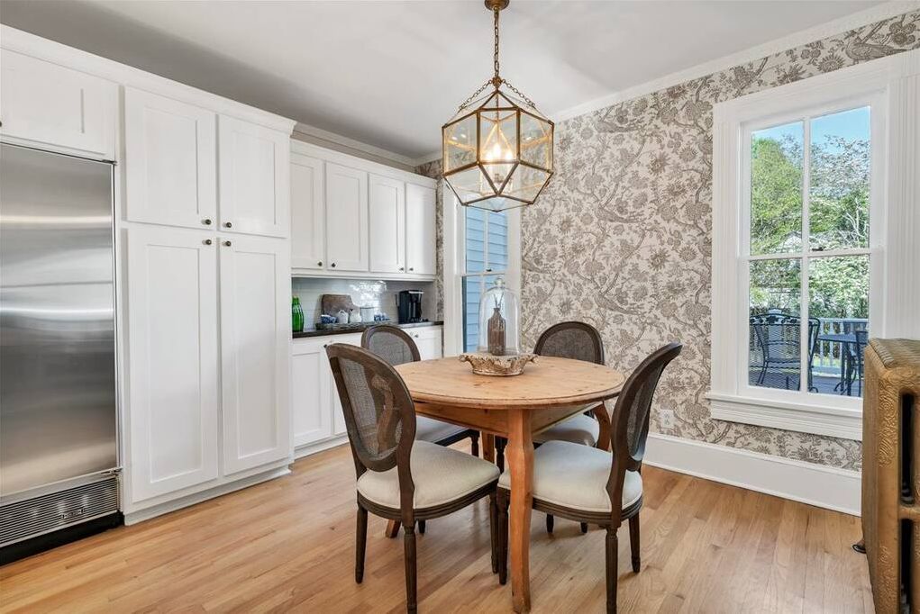 A kitchen with white cabinets, a stainless steel refrigerator, a wooden dining table with four chairs, and a patterned wallpaper.