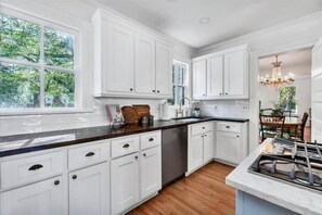 Una cocina con armarios blancos, encimera de madera y vista a árboles a través de ventanas amplias.
