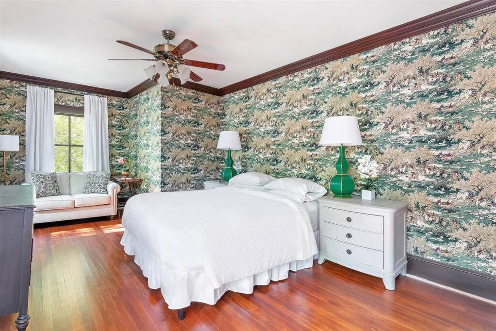 Une chambre avec un plancher en bois, un lit avec une literie blanche, une table de chevet avec une lampe verte et un ventilateur de plafond.