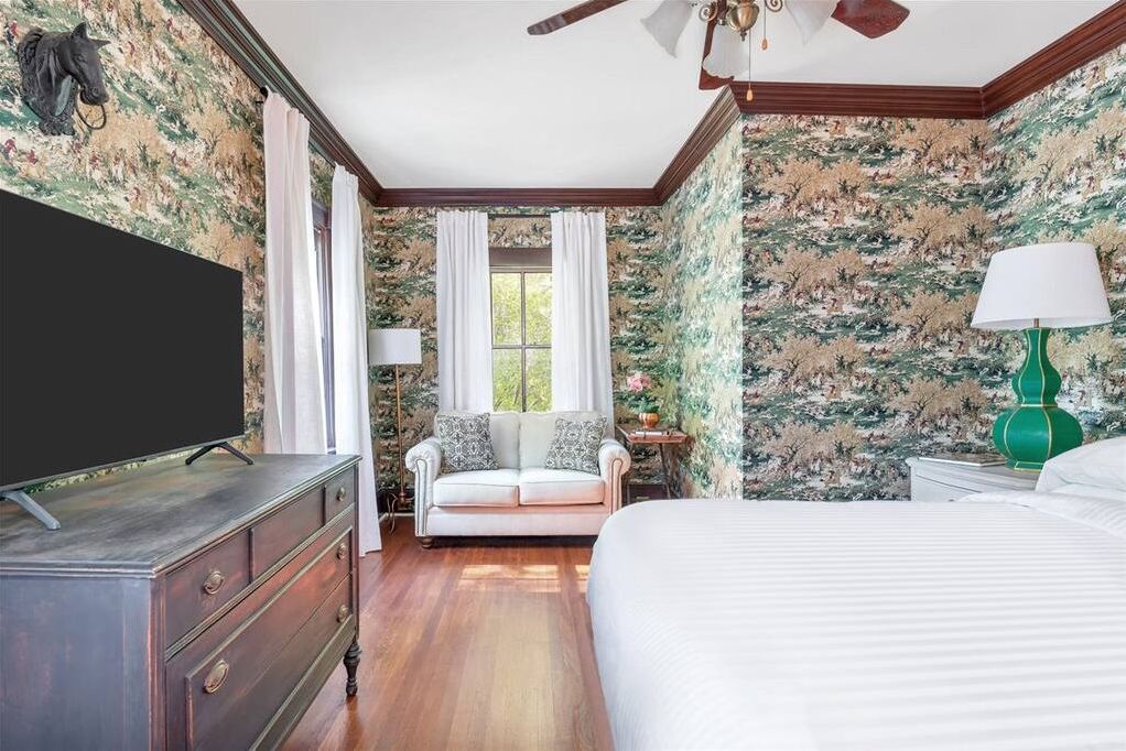 A bedroom with a bed, a dresser, a television, a window with curtains, and a wall with floral wallpaper.