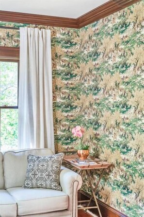 Une pièce avec un papier peint à motifs, une fenêtre avec des rideaux, un canapé agrémenté d’un coussin à motifs et une petite table sur laquelle est posé un vase.