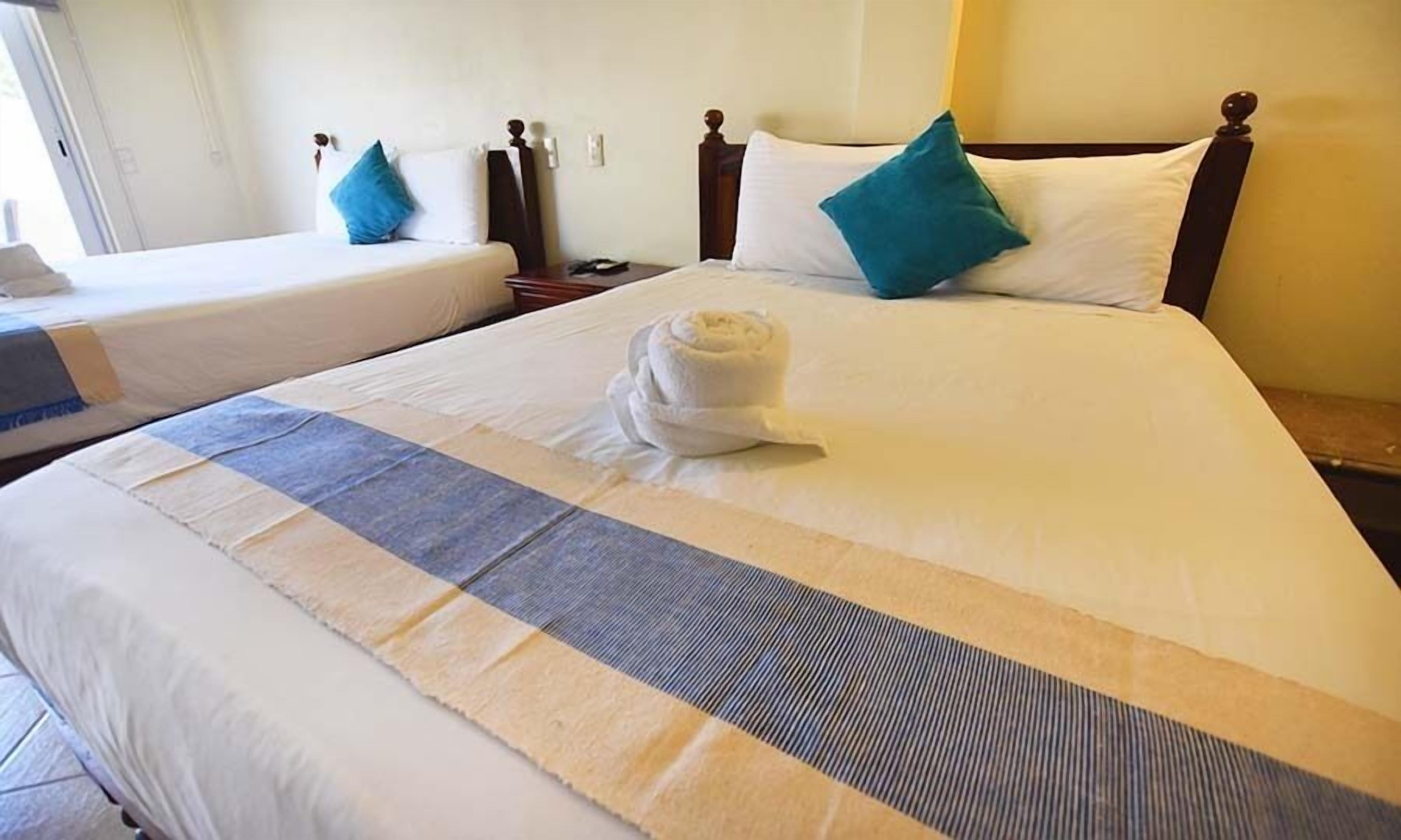 A hotel room with two single beds, a large bed with a rolled towel, and a striped area rug.