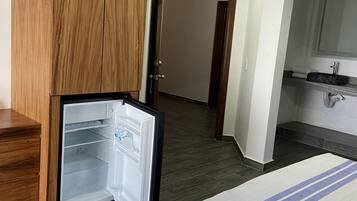 Habitación de hotel con un armario de madera, una cama con manta azul y blanca a rayas, un refrigerador pequeño, un baño con espejo y lavamanos, y piso de madera oscura.