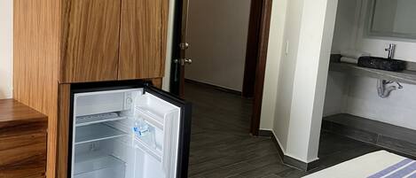A hotel room with a wooden wardrobe, a bed with a white and blue striped blanket, a small refrigerator, a bathroom with a mirror and sink, and a dark wooden floor.
