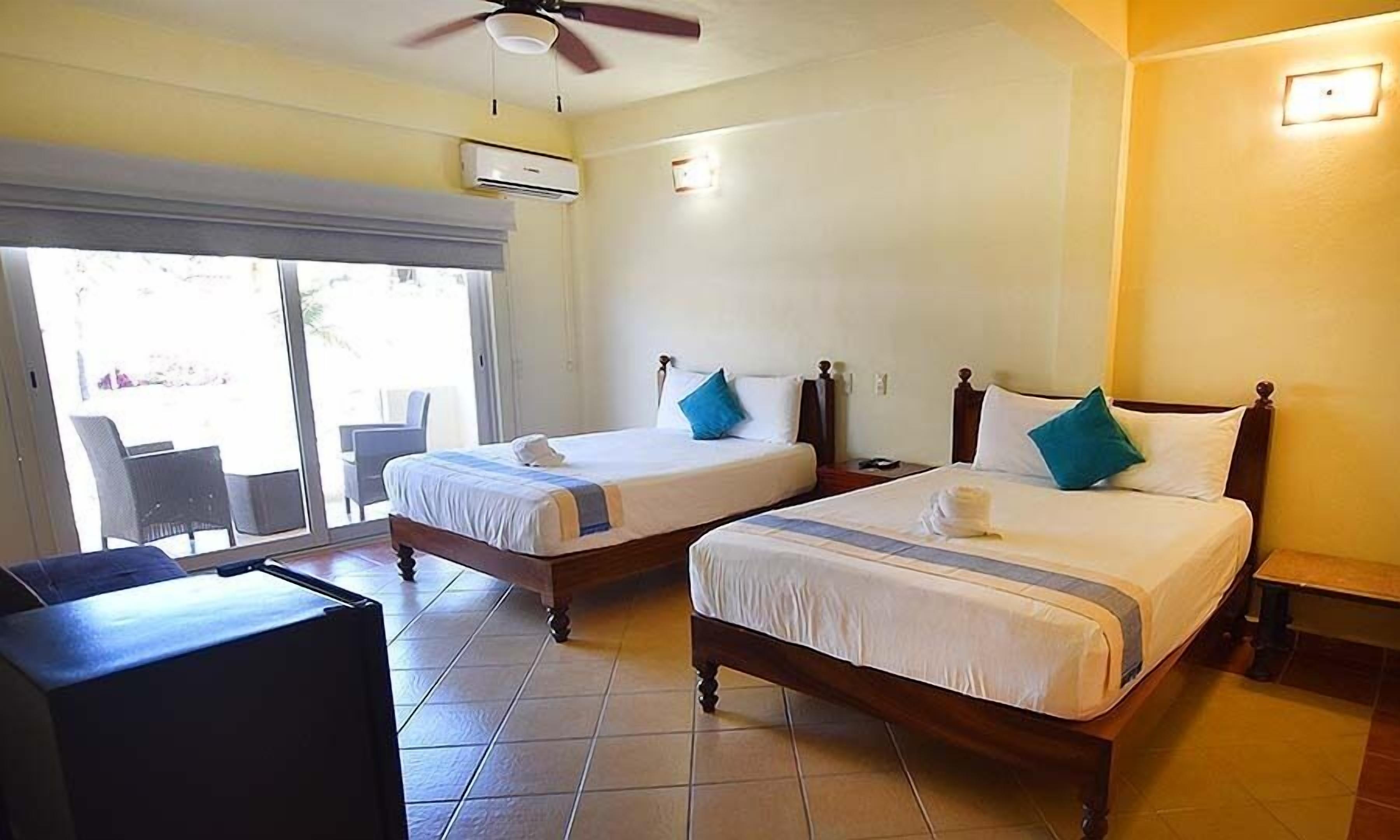 Une chambre d’hôtel avec deux lits, un ventilateur de plafond, un appareil de climatisation et une vue sur l’extérieur, accessible par une porte-fenêtre coulissante.