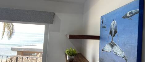 A room with a wooden shelf holding a potted plant, a painting of a whale, and a view of the ocean through a window.