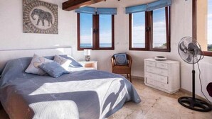 Un dormitorio con cama, mesita de noche, silla, cómoda y ventilador de pie.