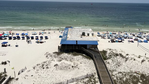 Une plage avec une promenade en bord de mer menant à un bâtiment doté d’un toit bleu, entouré de parasols et de chaises longues.