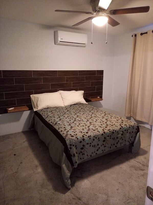 Un dormitorio con cama, ventilador de techo, unidad de aire acondicionado y ventana con cortinas.