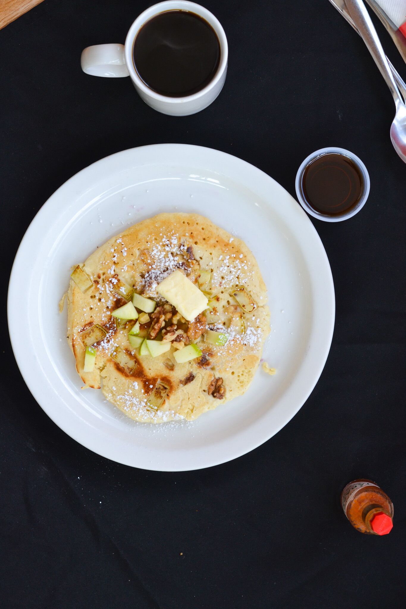 A plate of food with a cup of coffee and a small container of syrup on a black tablecloth.
