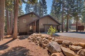 Situated in a forested area, this cabin features a front porch and a garage door, surrounded by a rock garden and lush trees.