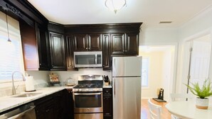Una cocina con armarios de madera oscura, electrodomésticos de acero inoxidable, lavavajillas y un comedor con mesa y sillas blancas.