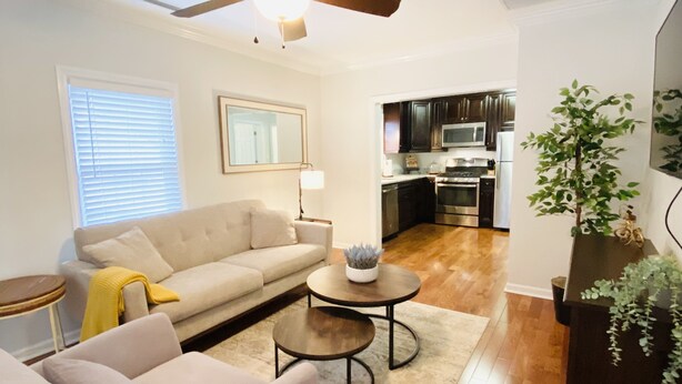 Una sala moderna con un sofá, dos sillones, una mesa de centro y un ventilador de techo. Se ve una cocina al fondo.