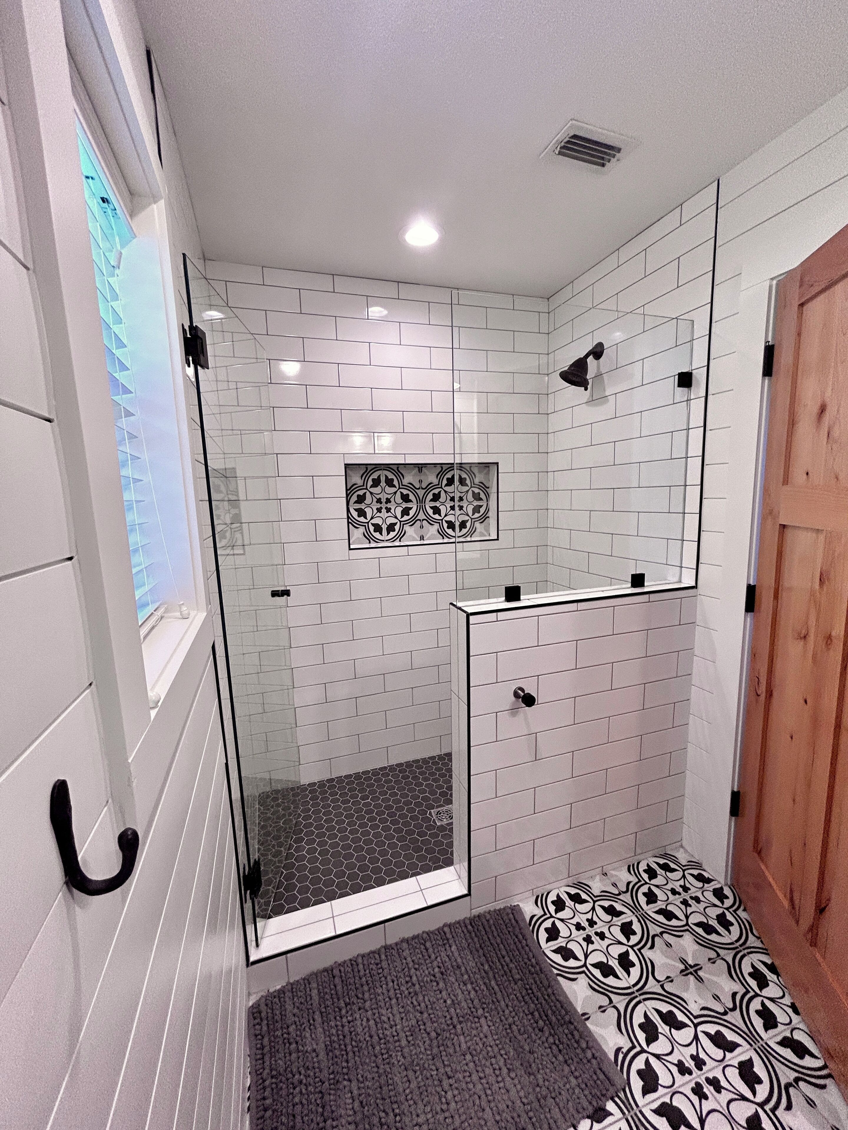 A modern bathroom with white subway tiles, a glass shower enclosure, a patterned floor, and a wooden door.