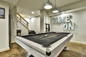 Une table de billard avec des queues et des billes, un escalier et un mur sur lequel est écrit « GAME ON ».