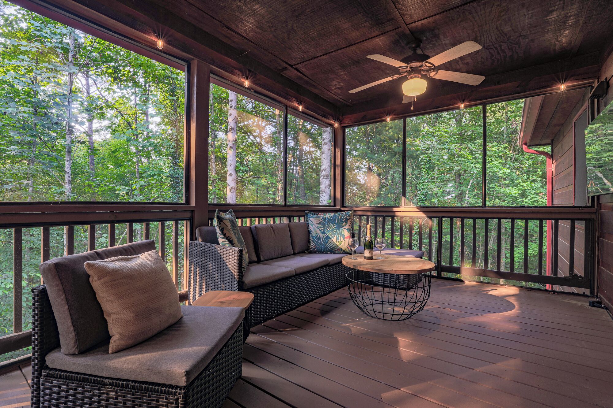 A covered outdoor seating area with a ceiling fan, wicker furniture, and a table with a bottle and glass.