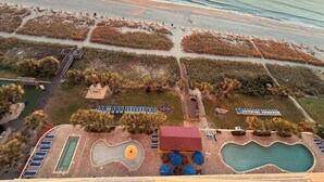 An aerial view of a beach resort with a swimming pool, lounge chairs, and a boardwalk.