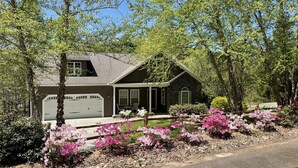 A two-story house with a garage, surrounded by lush greenery and flowering plants. - Peaceful Mountain Home: backs to a private fishing pond on two acres (Pickens)