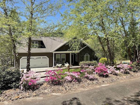 Une maison à deux étages avec un garage, entourée d’une végétation luxuriante et de plantes en fleurs.