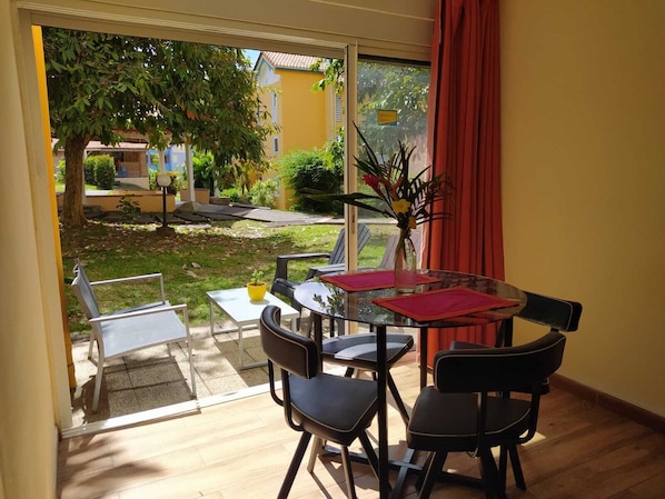 Une pièce dotée d’une porte vitrée donnant sur un patio. À l’intérieur, on trouve une table ronde avec un plateau en verre, entourée de quatre chaises. Un vase rempli de fleurs est posé sur la table.