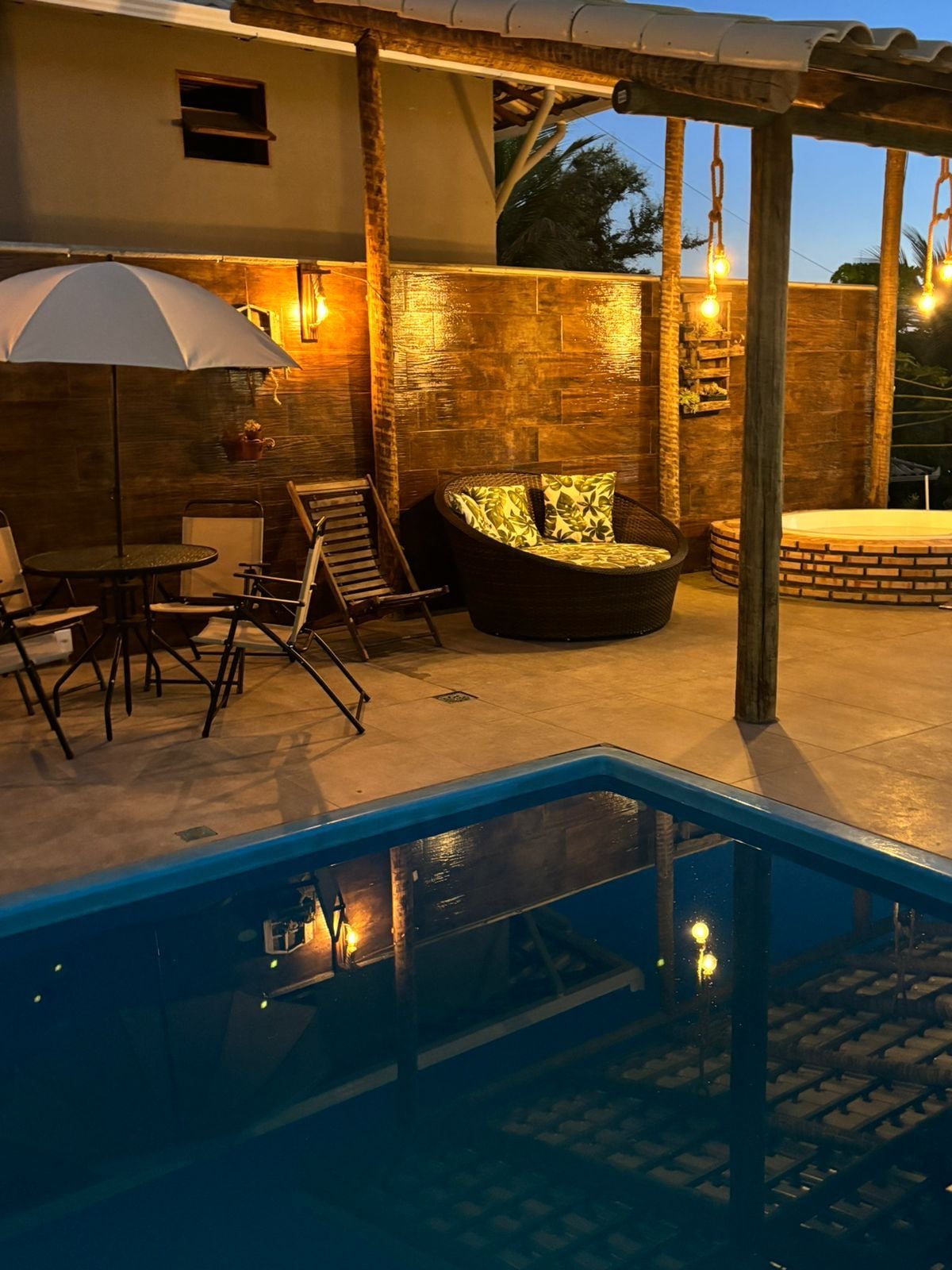 Een buitenzwembad met een ligbed, een tafel en een pergola met hangende lampen.