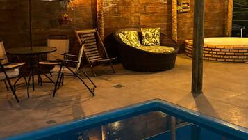 Área externa com piscina, espreguiçadeira, mesa e pergolado com luzes penduradas.