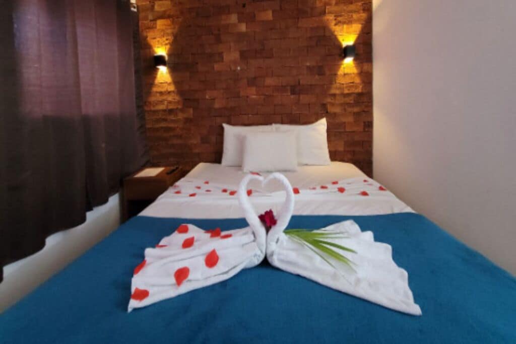 Habitación de hotel con pared de ladrillo, cama con ropa blanca y una toalla doblada en forma de corazón sobre la cama.