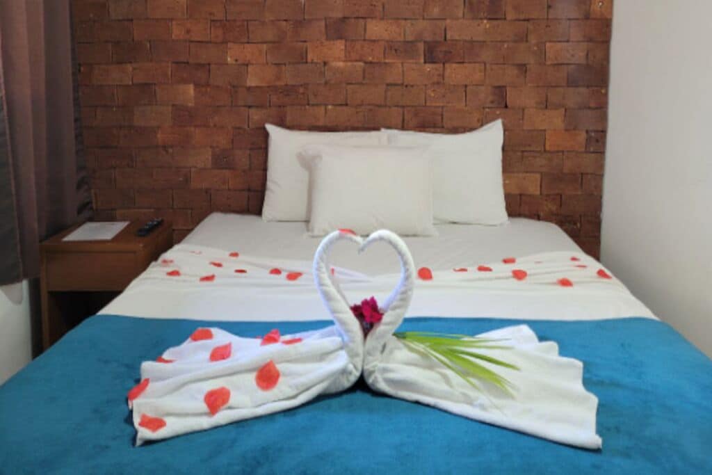 Quarto de hotel com cabeceira de tijolos, cama com roupa de cama branca e decoração de toalha em formato de coração sobre a cama.