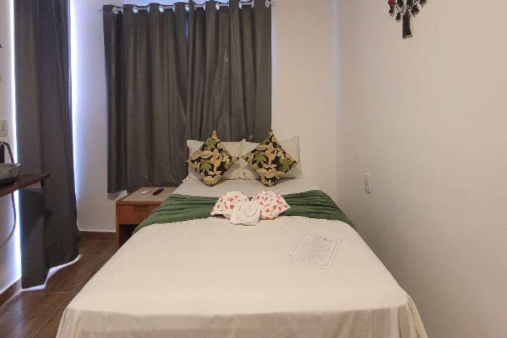 A bed with a white sheet, two pillows with a leaf pattern, a green blanket, and a small bedside table with a book.