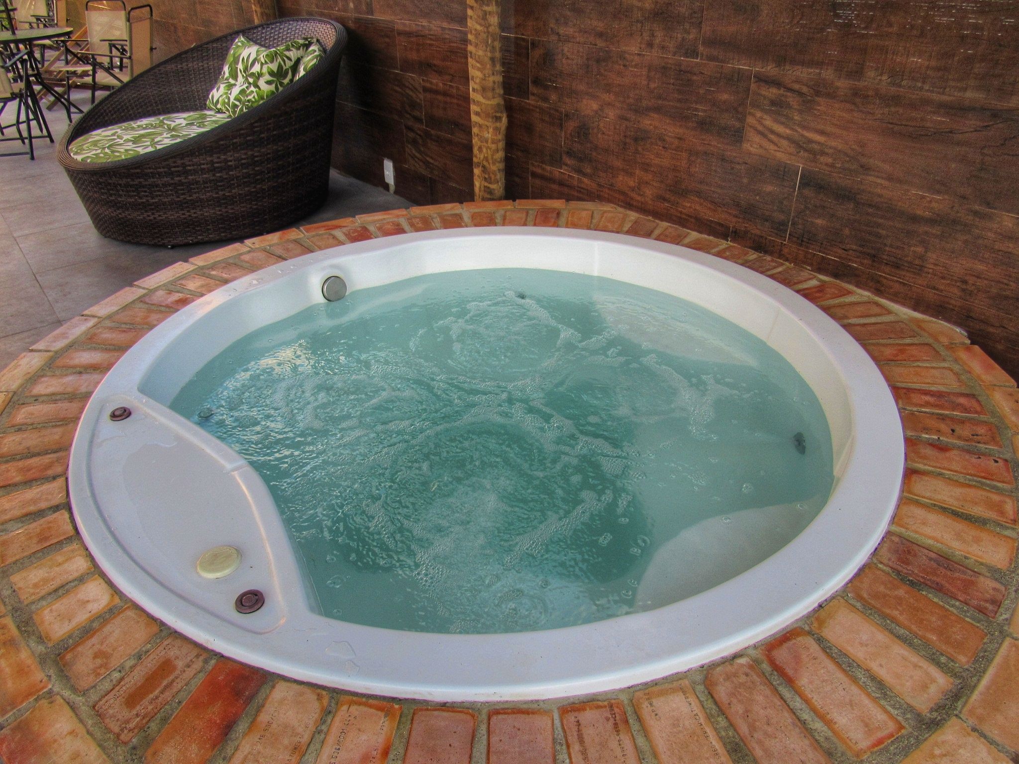 A hot tub with a brick border, surrounded by a patio area with a wicker chair and a table.