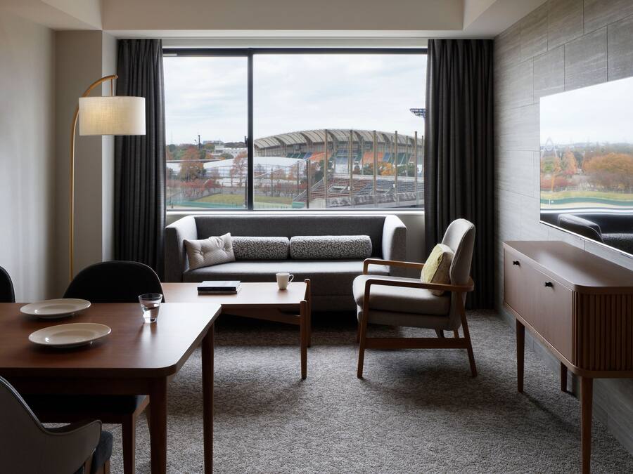 A hotel room with a sofa, armchair, dining table, and a large window offering a view of a stadium.