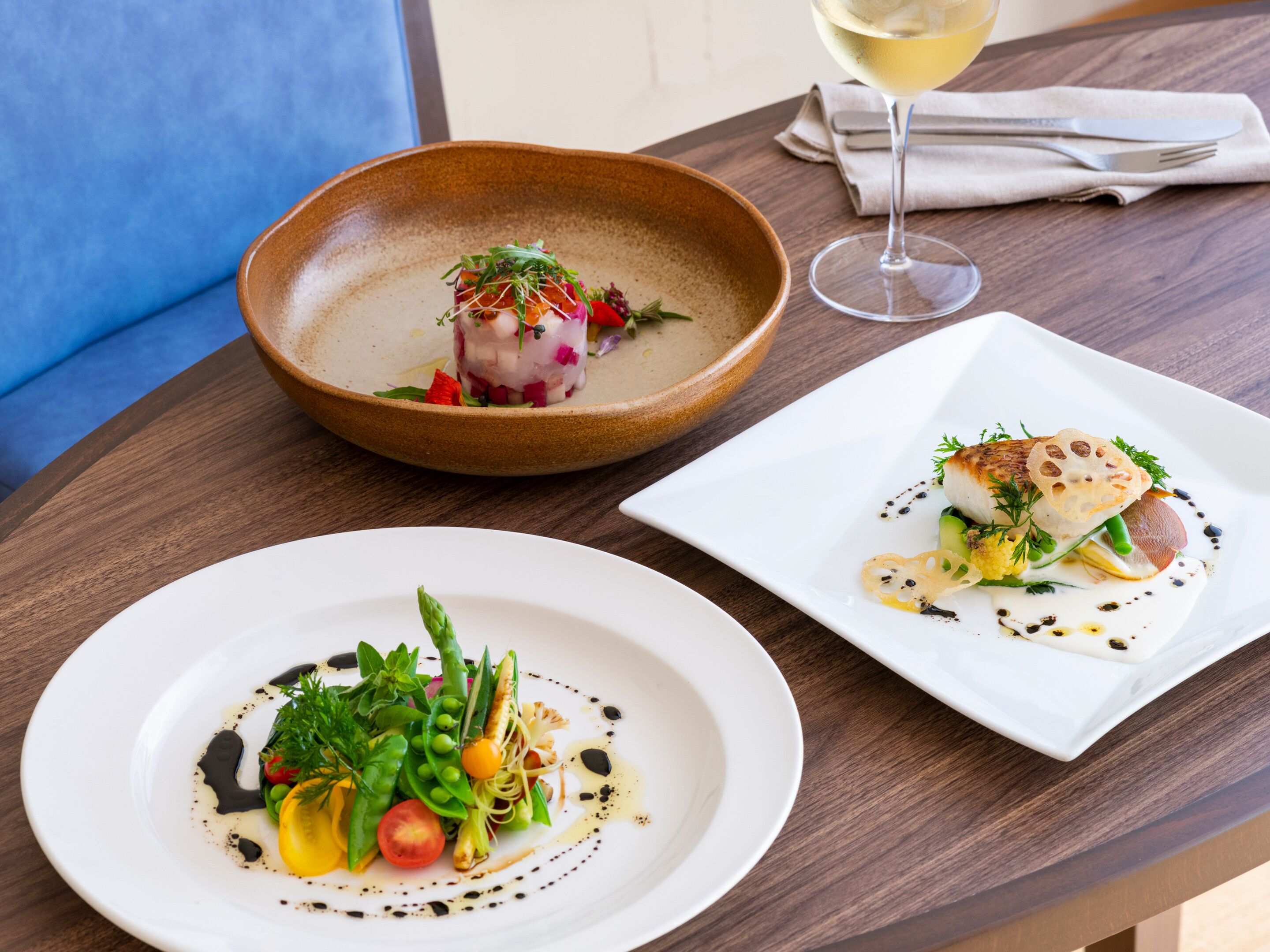Une table avec trois assiettes de nourriture, un verre de vin blanc et une serviette.