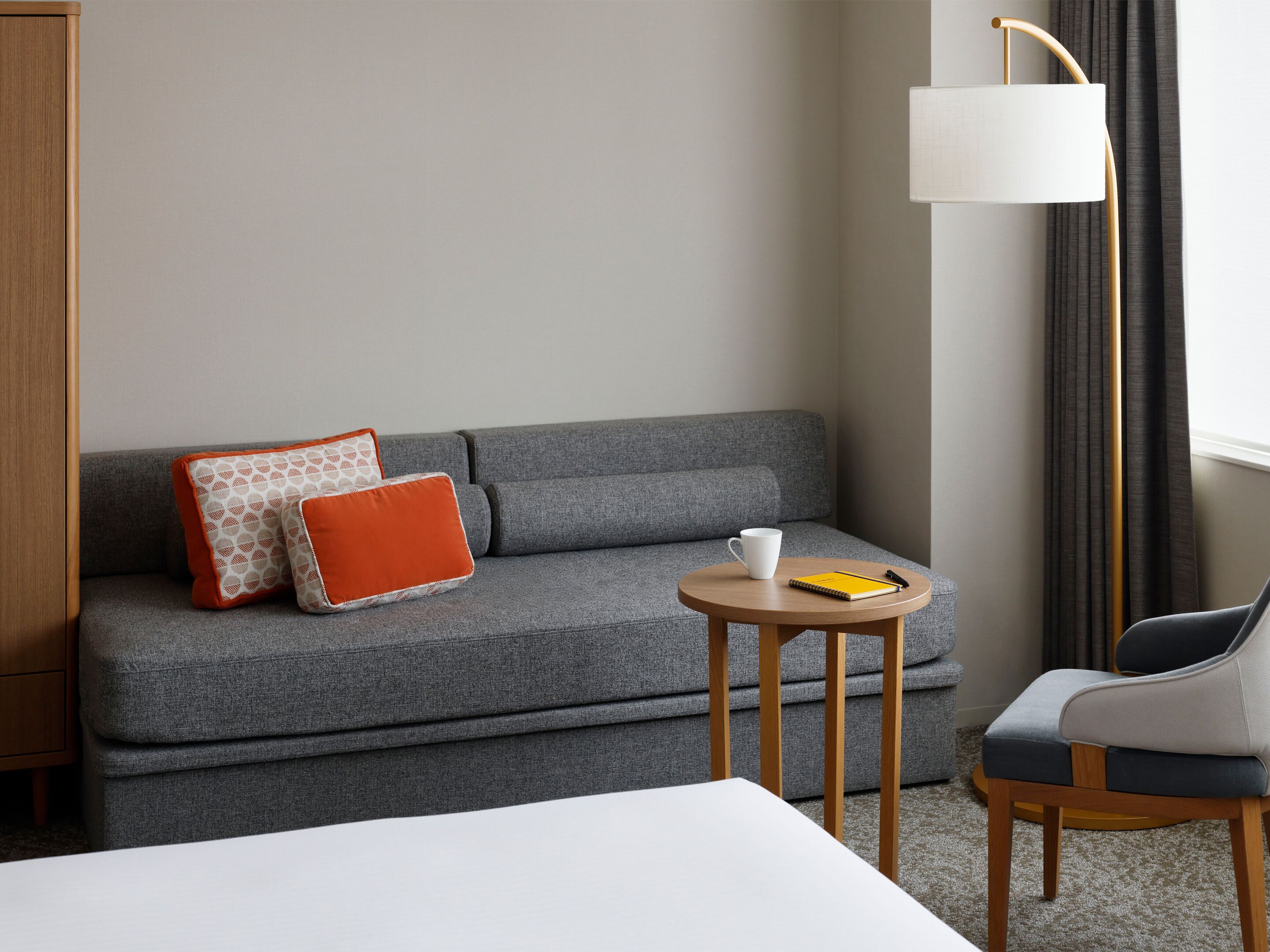 Habitación de hotel con un sofá gris, dos almohadas, una mesita redonda con una taza y un cuaderno, una silla y una lámpara de pie.