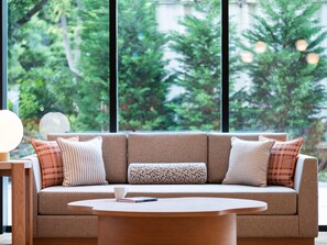 A modern living room with a sofa, patterned cushions, a round coffee table, and a view of greenery through large windows. - Mitsui Garden Hotel Kashiwanoha Park Side Chiba  (Kashiwa)