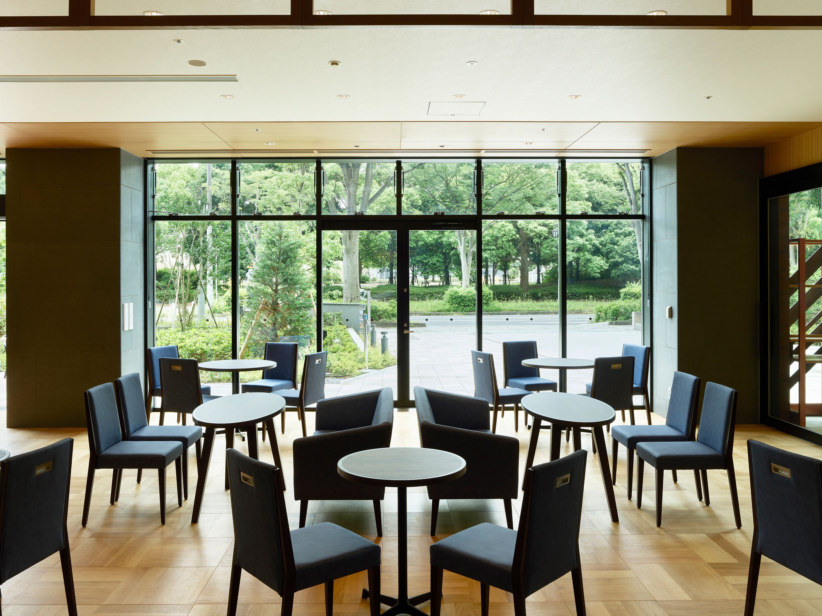 A modern indoor seating area with tables and chairs, large windows offering a view of a green outdoor space.