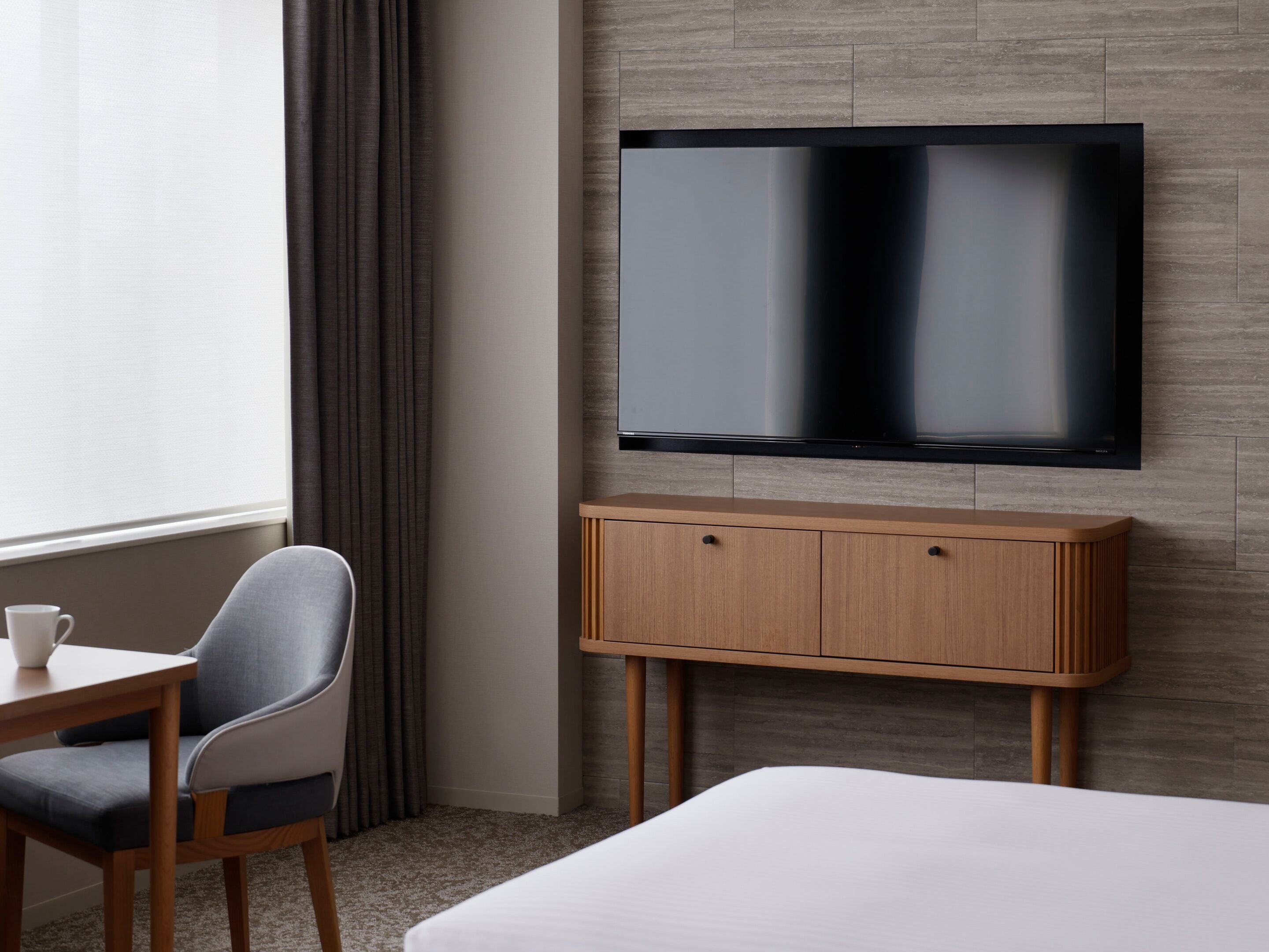 Une chambre d’hôtel avec un lit, un téléviseur fixé au mur, un meuble en bois placé sous le téléviseur, une chaise et une table avec une tasse.