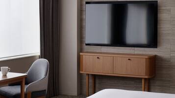 Habitación de hotel con cama, televisor montado en la pared, mueble bajo el televisor, una silla y una mesa con una taza.