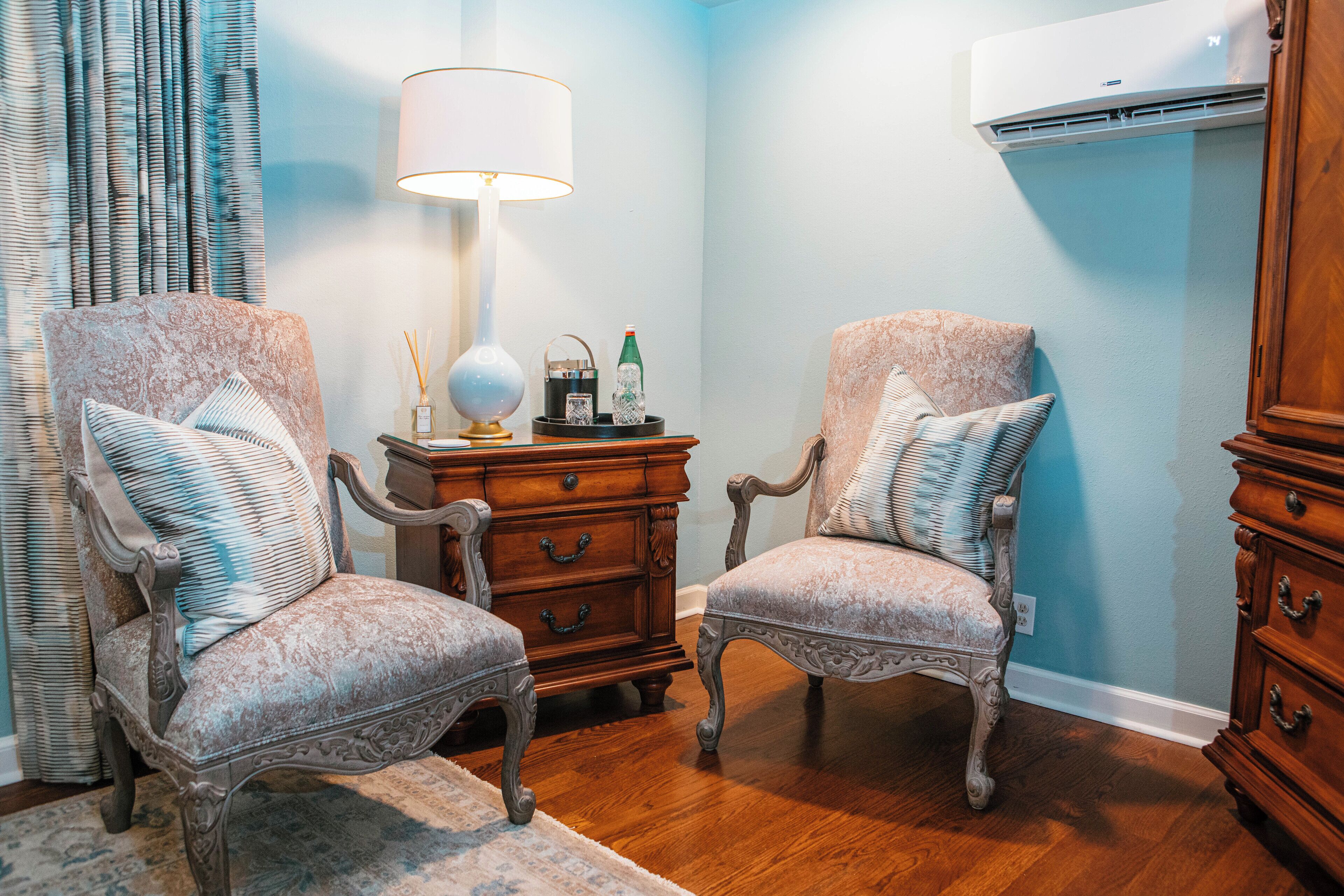 A room with two ornate chairs, a wooden side table with a lamp, and a wall-mounted air conditioner.