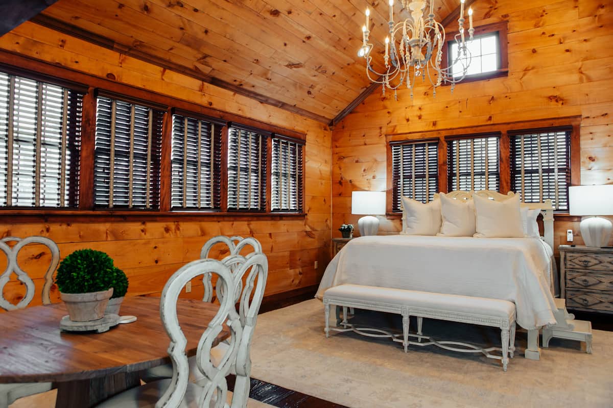 A log cabin bedroom with a large bed, white furniture, and multiple windows with blinds.
