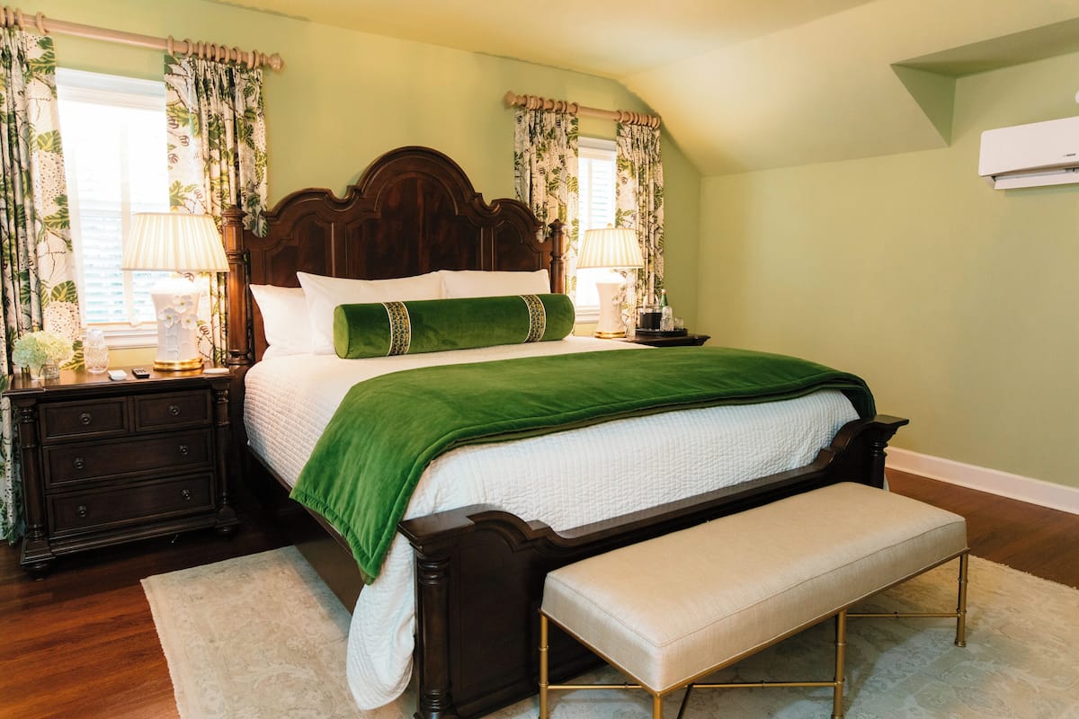 A bedroom with a large bed, a bench, a nightstand, a dresser, and a window with curtains.